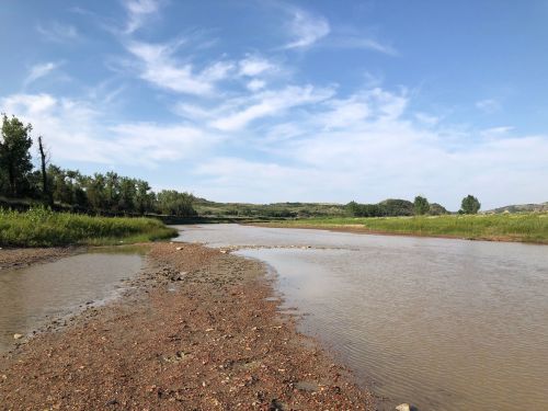 A serene river landscape with a sandy shore, surrounded by green trees and rolling hills under a blue sky with clouds.
