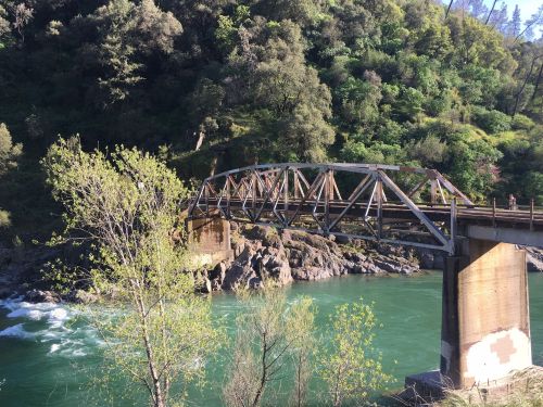 A wooden bridge spans a river surrounded by lush greenery and rocky banks under a clear blue sky.