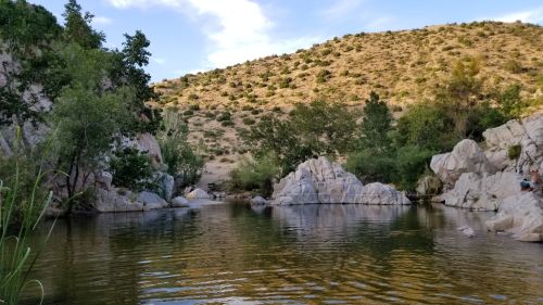 A serene landscape featuring a calm water body surrounded by rocky formations and lush greenery under a blue sky.