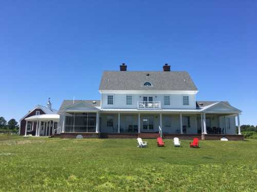 A large white house with a porch, surrounded by green grass and two red and white chairs in the foreground under a clear blue sky.