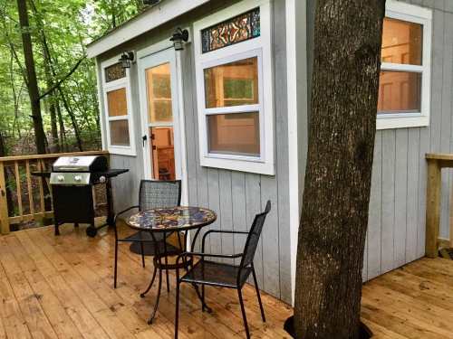 A cozy cabin with a wooden deck, featuring a grill and a table with chairs, surrounded by trees.