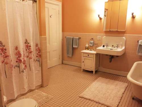 A cozy bathroom with peach walls, a floral shower curtain, a vintage sink, and a clawfoot tub on tiled flooring.