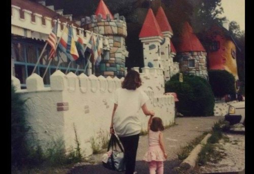 A woman and a young girl walk hand in hand past a colorful castle-themed building with flags.