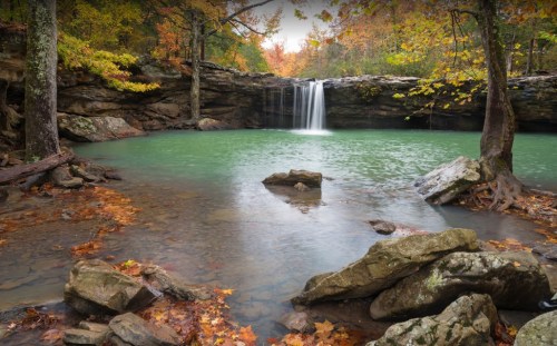 A serene waterfall cascades into a turquoise pool, surrounded by autumn foliage and rocky terrain.