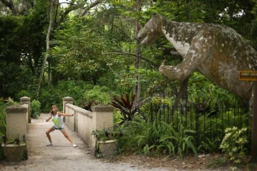 A girl poses playfully in front of a large dinosaur statue in a lush, green park setting.