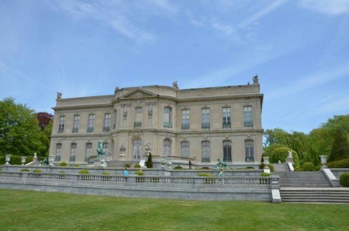 A grand mansion with ornate architecture, surrounded by manicured gardens and sculptures under a clear blue sky.