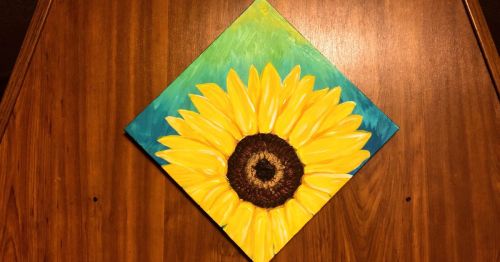 A vibrant painting of a sunflower with bright yellow petals against a blue-green background, displayed on a wooden surface.