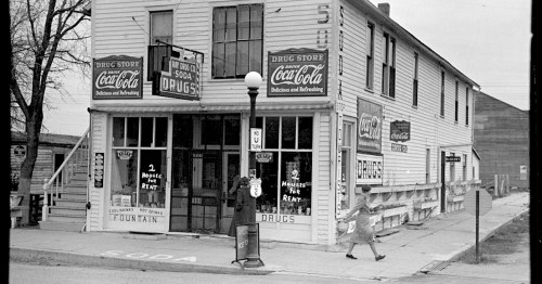 Historic drugstore with Coca-Cola signage, featuring a soda fountain and a woman walking by on the sidewalk.