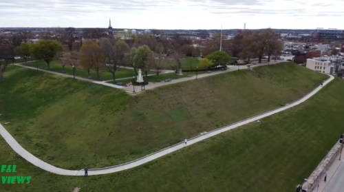 Aerial view of a park with winding paths, grassy hills, and trees, set against a cloudy sky and distant cityscape.