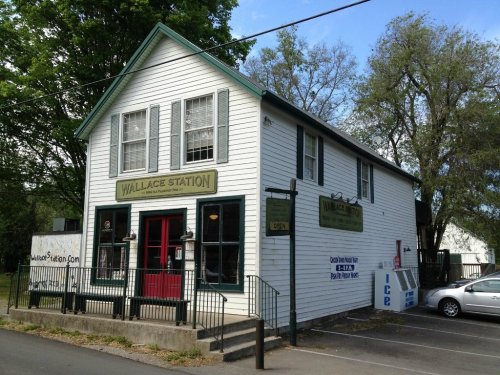A white, two-story building with green trim and signs reading "Wallace Station," surrounded by trees and a parking area.