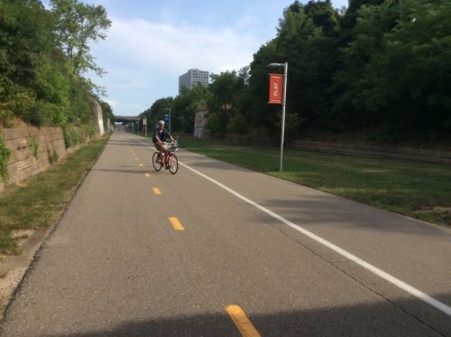 A cyclist rides on a paved path surrounded by greenery and buildings in the background.