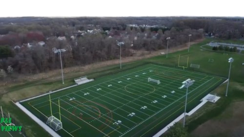 Aerial view of a soccer field with goalposts, surrounded by trees and a residential area in the background.