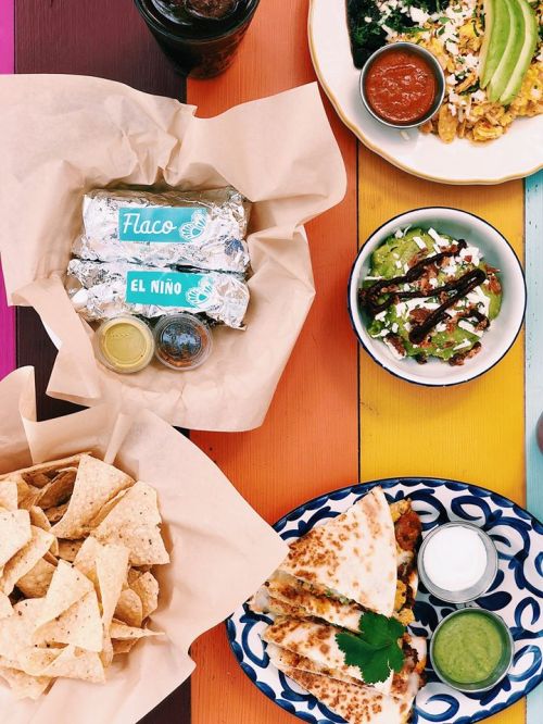 A colorful table spread featuring burritos, nachos, guacamole, and quesadillas with sauces and salsa.