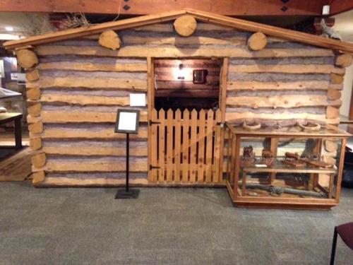 A rustic log cabin replica with a wooden door and a display case nearby, set in a museum environment.