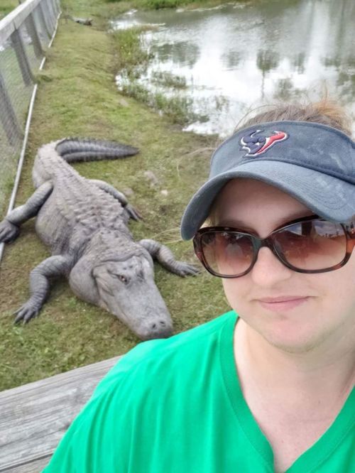A woman in sunglasses and a cap poses for a selfie with a large alligator resting nearby.