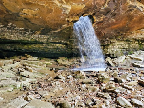 A waterfall cascades from a rocky overhang into a stony, forested area below.