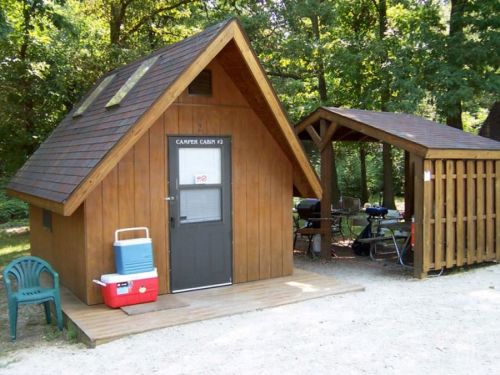 A small wooden cabin with a sloped roof, next to a covered picnic area and a cooler on the ground. Surrounded by trees.