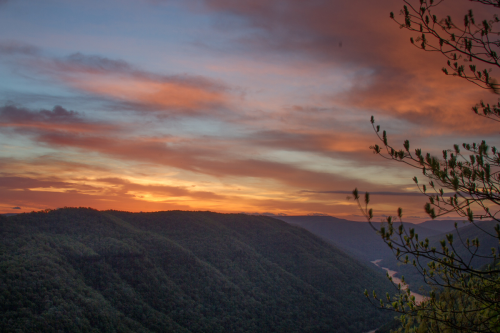 A vibrant sunset over rolling green mountains, with colorful clouds and a hint of a river winding through the valley.