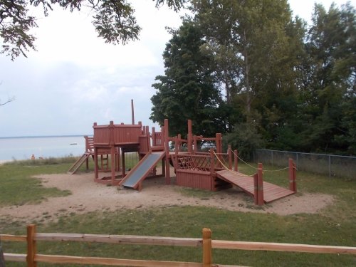 A wooden playground structure with slides and climbing areas, surrounded by grass and trees near a lake.