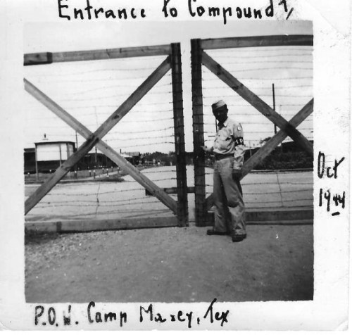 Black and white photo of a soldier standing by a barbed wire gate labeled "Entrance to Compound 7, P.O.W. Camp Marcy, Tex."