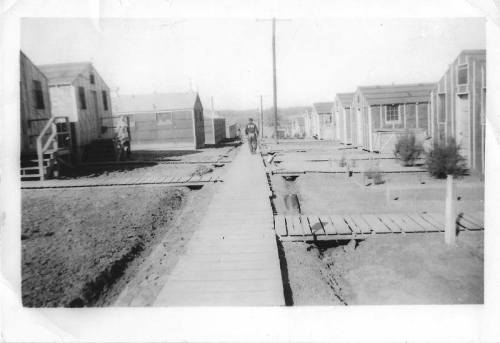 Black and white photo of a dirt path lined with small wooden houses in a sparse settlement.