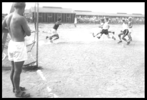 A black and white photo of a soccer game, with players running towards the goal and a spectator watching from the side.