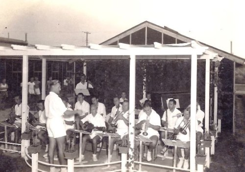 A vintage black-and-white photo of a band performing outdoors, with musicians in white uniforms and a conductor.