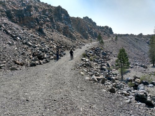 Two hikers walk along a rocky trail surrounded by steep, rugged terrain and sparse trees.