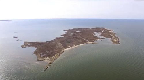 Aerial view of a small, rugged island surrounded by calm waters, featuring sandy shores and rocky outcrops.