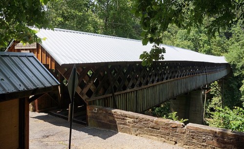 A covered bridge with a white roof spans a lush green landscape, surrounded by trees and stone walls.