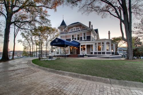 A large, elegant house with a turret, surrounded by trees and a paved driveway, at sunset.