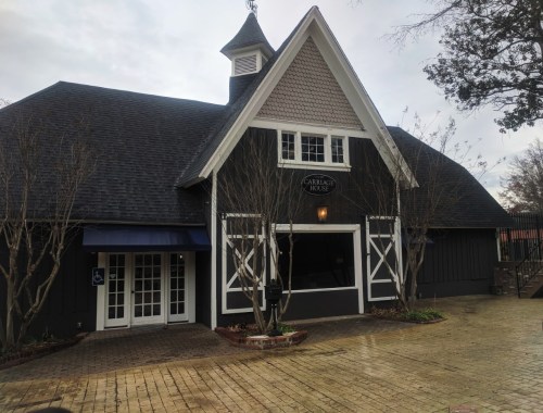 A large, dark wooden building with a peaked roof, featuring white trim and a sign that reads "Carriage House."