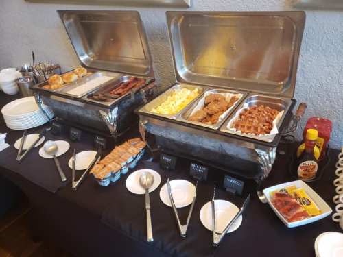 A buffet setup with two chafing dishes containing eggs, bacon, chicken, waffles, and various condiments on a table.