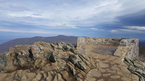 A rocky overlook with a stone wall, offering a panoramic view of distant mountains under a cloudy sky.