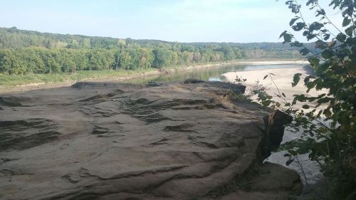 A rocky cliff overlooks a calm river surrounded by lush green trees and a clear blue sky.