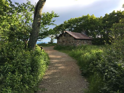 A gravel path leads to a rustic stone house surrounded by lush greenery and trees under a cloudy sky.