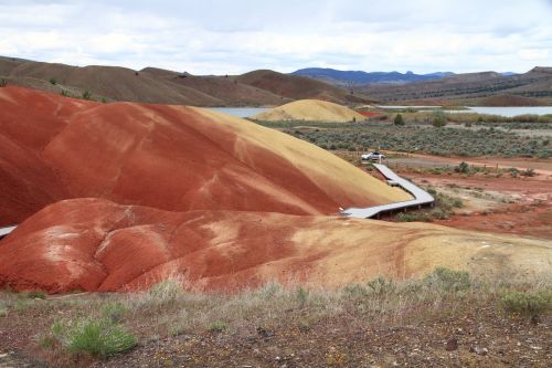 Colorful hills of red, yellow, and brown with a winding path and a distant lake under a cloudy sky.