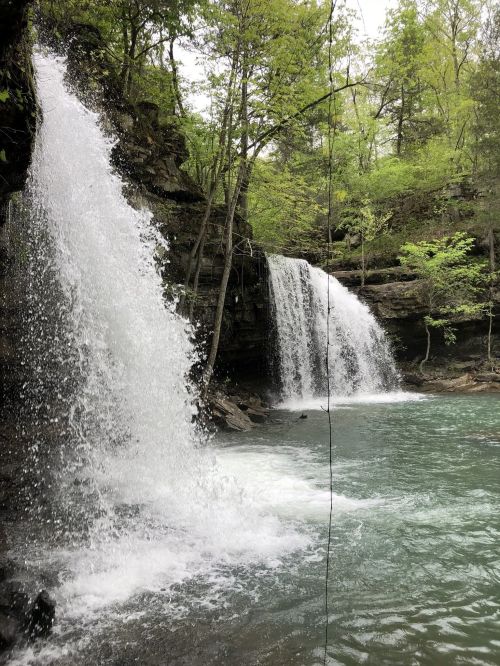 Two waterfalls cascade into a serene turquoise pool, surrounded by lush green trees and rocky terrain.
