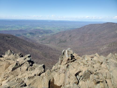 A panoramic view of a valley surrounded by mountains, with rocky foreground and clear blue sky above.