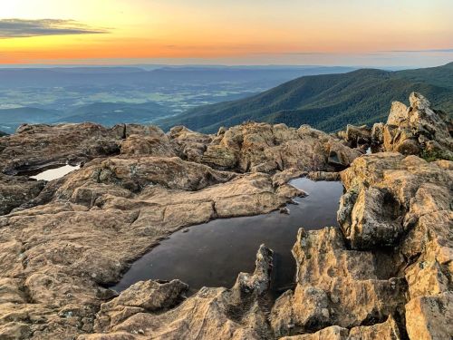 Sunset over rocky terrain with small pools of water, surrounded by rolling hills and distant mountains.