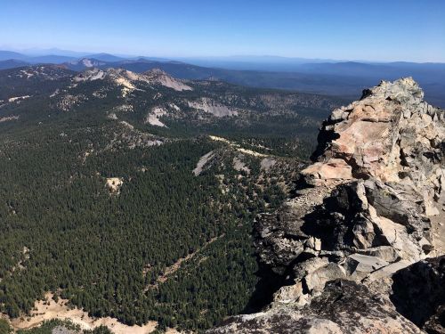 A panoramic view of rugged mountains and dense forests under a clear blue sky. Rocky outcrop in the foreground.