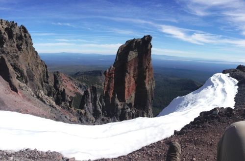 A panoramic view from a mountain peak, featuring rocky cliffs, a snow patch, and a vast landscape below.