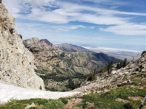A panoramic view of rugged mountains and valleys under a blue sky with scattered clouds. Snow patches and greenery visible.