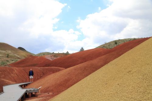 A person stands on a wooden path surrounded by colorful, rolling hills of red and yellow earth under a cloudy sky.