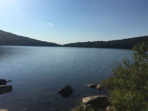 A serene lake surrounded by green hills under a clear blue sky, with rocks in the foreground.
