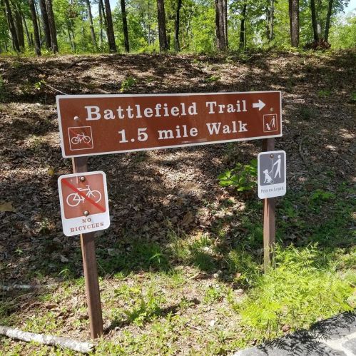 Sign for Battlefield Trail indicating a 1.5-mile walking path, with no bicycles allowed. Surrounded by trees.