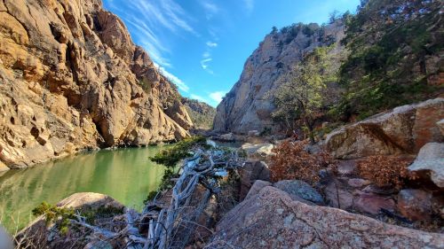 A serene river flows between towering rock formations, surrounded by trees and a clear blue sky.