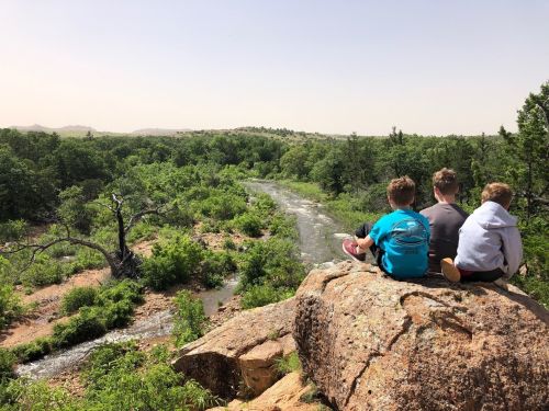 Three children sit on a rock overlooking a lush green landscape and a winding river under a clear sky.