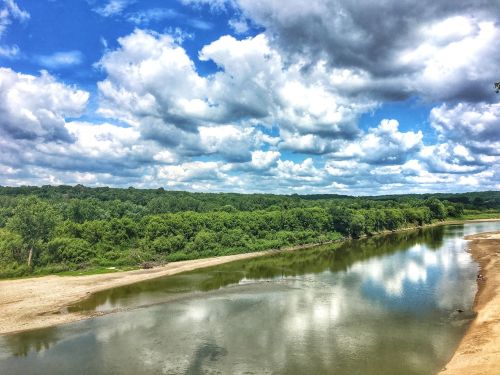 A serene river flows through lush greenery under a sky filled with fluffy clouds.