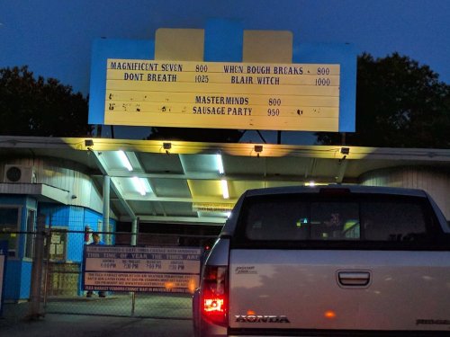 A drive-in theater marquee displays movie titles and showtimes at dusk, with a truck parked in front.